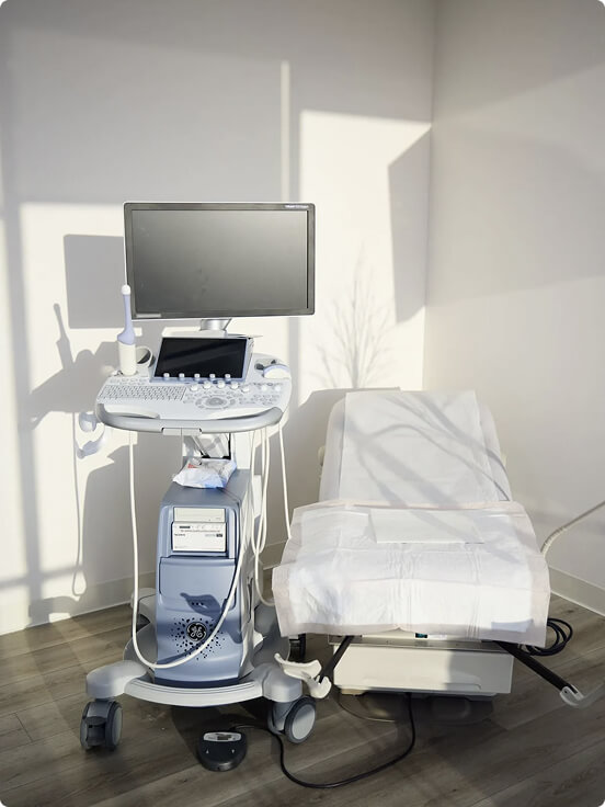 Ultrasound machine sits beside an examination bed in a well-lit medical room, with shadows cast on the walls, creating a clean and clinical environment.