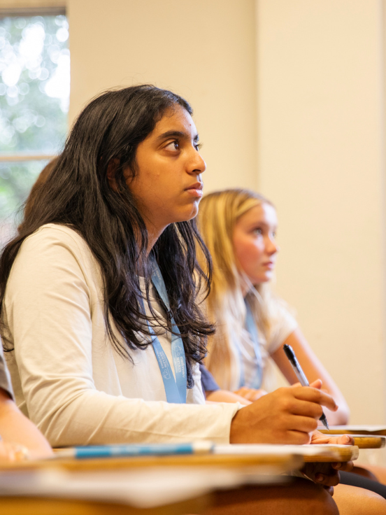 Two students sit and write attentively in a classroom, with desks, notebooks, and a window in the background providing a focused learning environment.