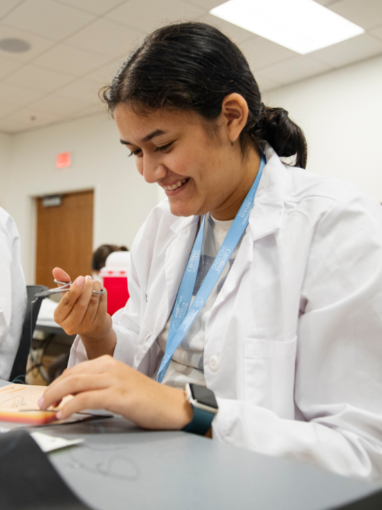 A student interested in medicine in a lab coat smiles while using a tool to work on a model. They sit at a table in a bright room with ceiling lights.