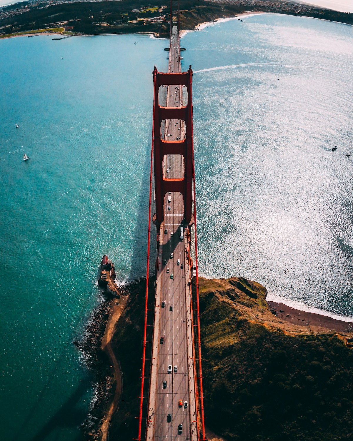 Red suspension bridge spans across a body of shimmering water. Vehicles travel along the bridge, with green hills and a distant shoreline in the background.