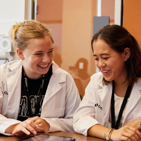 Two students in white lab coats are seated at a table, smiling and engaged in conversation. They appear to be in a bright room with warm-toned walls.